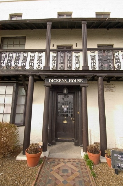 Looking through black painted wooden pillars around door entrance holding up balcony above. Dickens House written in white text above door and can see windows to left side and above. Building painted white. Mosaic tiled path leading to door